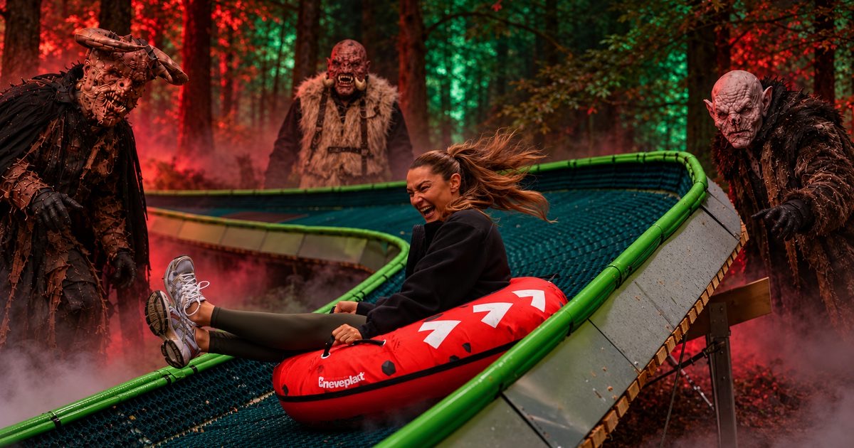A woman laughing as she slides down a forest tubing run at night in a red tube, while three costumed monsters watch from the trees.
